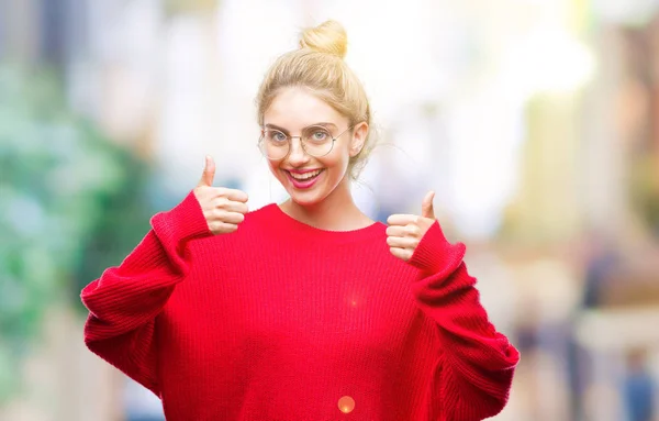 Young beautiful blonde woman wearing red sweater and glasses over isolated background success sign doing positive gesture with hand, thumbs up smiling and happy. Looking at the camera with cheerful expression, winner gesture.