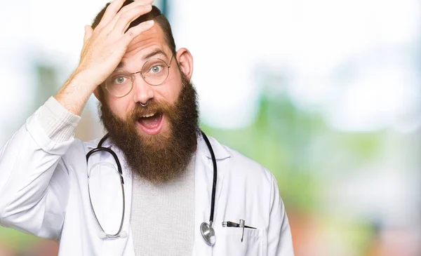 Young blond doctor man with beard wearing medical coat surprised with hand on head for mistake, remember error. Forgot, bad memory concept.