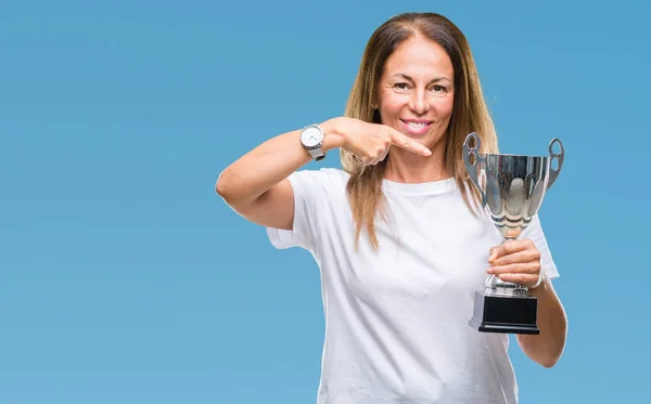 Middle age hispanic winner woman celebrating award holding trophy over isolated background very happy pointing with hand and finger