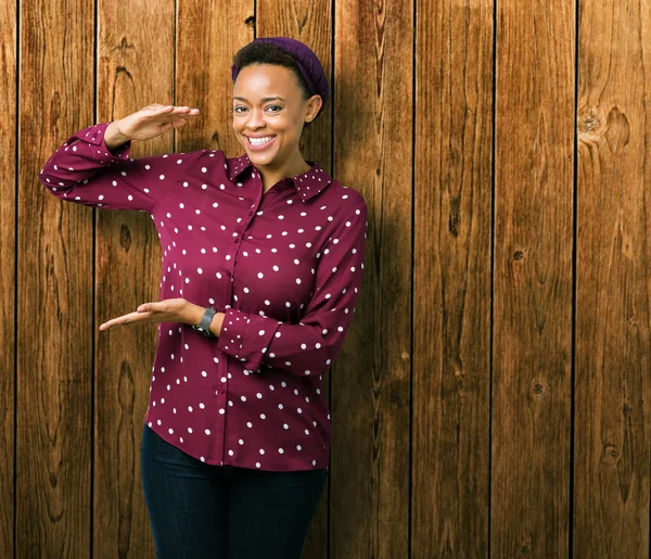 Beautiful young african american woman wearing head scarf over isolated background gesturing with hands showing big and large size sign, measure symbol. Smiling looking at the camera. Measuring concept.