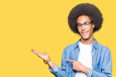 Hayret ve el ile sunulması ve parmak ile işaret ederken kameraya gülümseyen gözlüklü afro saçlı genç Afro-Amerikan adam.