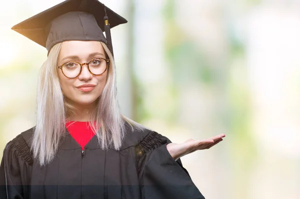 Young blonde woman wearing graduate uniform over isolated background smiling cheerful presenting and pointing with palm of hand looking at the camera.