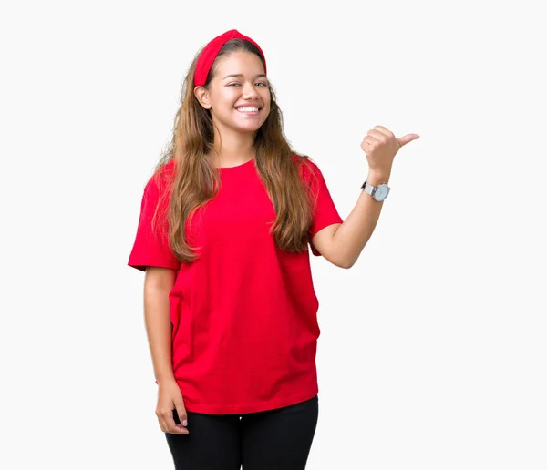 年轻美丽的黑发女人穿着红色的 T恤在孤立的背景微笑着与快乐的脸看 用拇指指着一边 — 图库照片