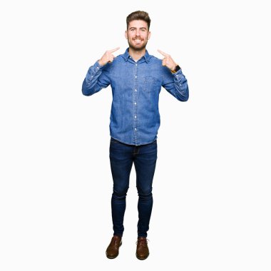 Kendinden emin gösterilen ve işaret parmakları diş ve ağız ile gülümseyen rahat kot gömlek giymiş genç yakışıklı sarışın adam. Sağlık kavramı.