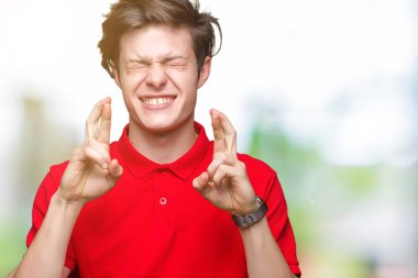 Umut ve gözleri kapalı geçiş parmaklarıyla gülümseyen izole arka plan üzerinde kırmızı t-shirt giyen genç yakışıklı adam. Şans ve batıl kavramı.