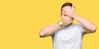 Yakışıklı adam giyiyor rahat beyaz t-shirt kapsayan gözleri ve şaşırttı ve şok ellerle ağız. Duygu gizleme