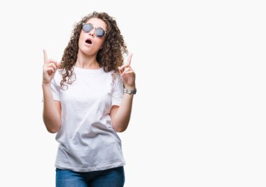 Güzel esmer kıvırcık saçlı Genç kız izole arka plan üzerinde güneş gözlüğü takıyor hayret ve yukarıya bakıp parmakları ile işaret şaşırttı ve silah kaldırdı.