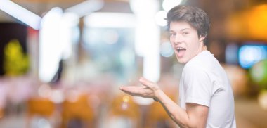 Yakışıklı delikanlı üzerinde rahat beyaz t-shirt giyen izole arka plan el ve reklam mutlu ve kendinden emin gülümseyen sunulması avuç, yanında işaret