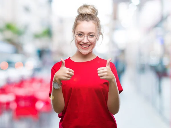 Young blonde woman wearing glasses over isolated background success sign doing positive gesture with hand, thumbs up smiling and happy. Looking at the camera with cheerful expression, winner gesture.