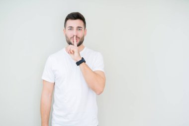 Dudaklarında parmak ile sessiz olmanı izole arka plan üzerinde rahat beyaz t-shirt giyen genç yakışıklı adam. Sessizlik ve gizli kavramı.