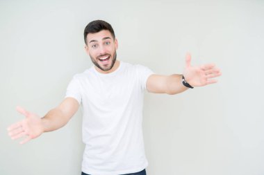 Sarılmak için kollarını açarak gülümseyerek kameraya bakarak izole arka plan üzerinde rahat beyaz t-shirt giyen genç yakışıklı adam. Neşeli ifade kucaklayan mutluluk.
