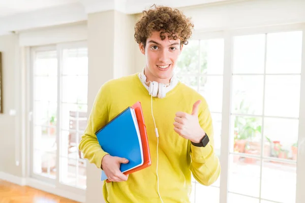 Young student man wearing headphones and holding notebooks happy with big smile doing ok sign, thumb up with fingers, excellent sign