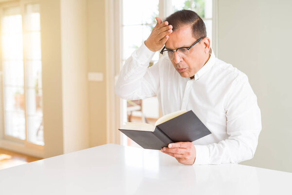 Middle age man reading a book at home wearing glasses stressed with hand on head, shocked with shame and surprise face, angry and frustrated. Fear and upset for mistake.