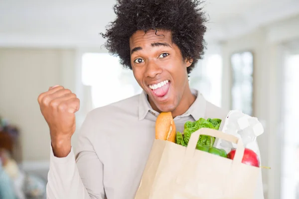 African American man holding groceries bag with fresh vegetables at home screaming proud and celebrating victory and success very excited, cheering emotion