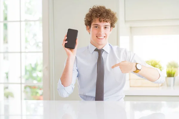 Young business man showing smartphone screen at the office with surprise face pointing finger to himself