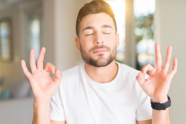 Casual beyaz t-shirt evde dinlenmek giydiği ve gözleri ile gülümseyen yakışıklı delikanlı parmak ile meditasyon jest yapıyor kapalı. Yoga kavramı.