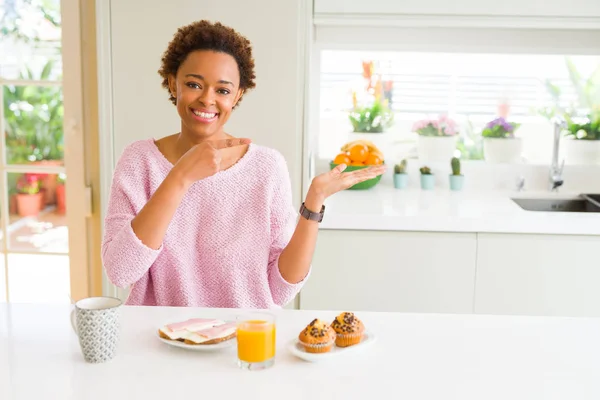Young african american woman eating breaksfast in the morning at home amazed and smiling to the camera while presenting with hand and pointing with finger.