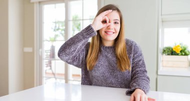 Güzel genç kadın evde gülümseyen, parmakları ile mutlu yüz üzerinden seyir göz el ile ok jest yapıyor.