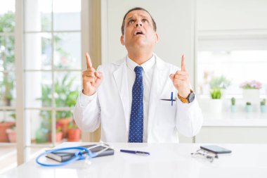 Orta yaş doktor adam klinikte tıbbi bir palto giyiyor hayrete ve yukarıya bakıyor ve parmakları ile işaret ve silah kaldırdı şaşırttı.