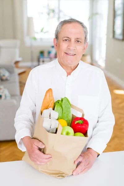 Handsome senior man holding paper bag full of fresh groceries and smiling at home