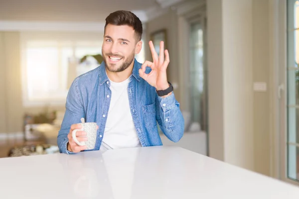 Young handsome man drinking a cup of coffee at home doing ok sign with fingers, excellent symbol