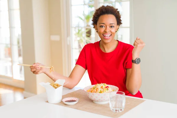 Young african american woman with afro hair eating asian food at home screaming proud and celebrating victory and success very excited, cheering emotion