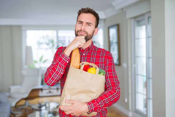 Handsome man holding groceries bag serious face thinking about question, very confused idea