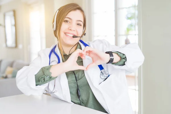 Beautiful young doctor woman wearing headset smiling in love showing heart symbol and shape with hands. Romantic concept.
