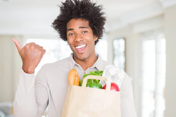 アフリカ系アメリカ人の家で新鮮な野菜の食料品袋を抱えて指していると幸せそうな顔を笑顔で側まで親指で示す — ストック写真