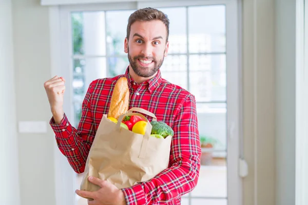 Handsome man holding groceries bag screaming proud and celebrating victory and success very excited, cheering emotion