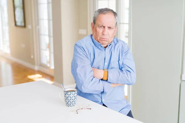 Handsome senior man drinking a cup of coffee at home skeptic and nervous, disapproving expression on face with crossed arms. Negative person.