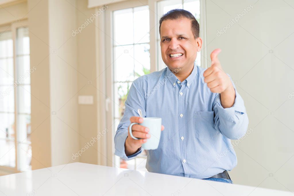 Middle age man drinking coffee in the morning at home happy with big smile doing ok sign, thumb up with fingers, excellent sign