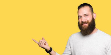 Kameraya bakarak yan el ve parmak ile işaret yüzünde kocaman bir gülümseme ile rahat kazak giyen genç sarışın adam.