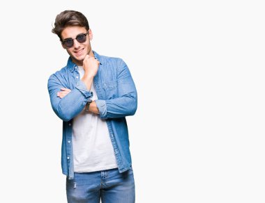 Genç yakışıklı adam emin çapraz kollar ve çeneme kaldırdı el ile gülümseme ile kameraya bakarak izole arka plan üzerinde güneş gözlüğü takıyor. Pozitif düşünce.