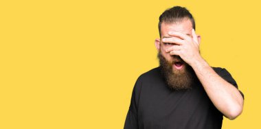 Rahat t-shirt yüz ve gözler el ile kapsayan şok bakmak mahcup ifadeyle parmaklarımın arasından seyir giyen genç sarışın adam.