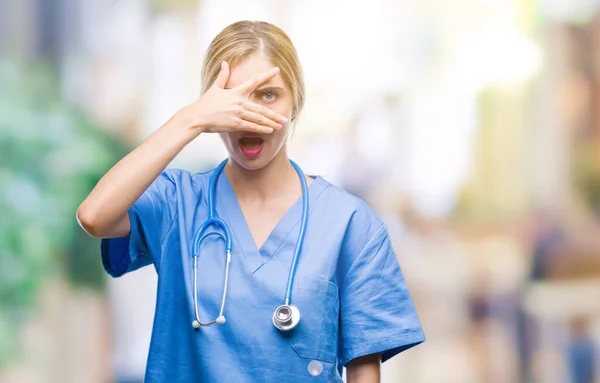 Young beautiful blonde doctor surgeon nurse woman over isolated background peeking in shock covering face and eyes with hand, looking through fingers with embarrassed expression.