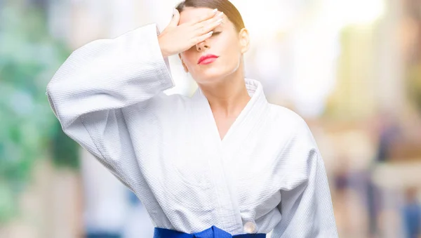 Young beautiful woman wearing karate kimono uniform over blurred background smiling and laughing with hand on face covering eyes for surprise. Blind concept.