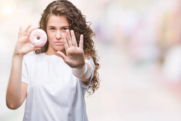 防衛ジェスチャー 深刻な自信を持って式に一時停止の標識を行うオープンの手で隔離された背景にドーナツを食べる若いブルネットの少女 — ストック写真