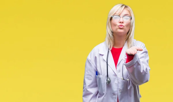 Young beautiful blonde doctor woman wearing medical uniform over isolated background looking at the camera blowing a kiss with hand on air being lovely and sexy. Love expression.