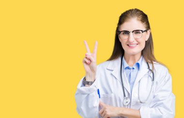 Orta yaş olgun doktor kadın mutlu yüz zafer işareti yaparken kameraya göz kırpıyor ile gülümseyen izole arka plan üzerinde tıbbi bir palto giyiyor. İki numaralı.
