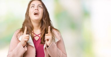 Artı boyutu genç kadın güzel izole arka plan üzerinde kış palto giyiyor hayret ve yukarıya bakıyor ve parmakları ile işaret ve silah kaldırdı şaşırttı.