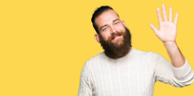 Kış kazak Waiving selam söyleyerek mutlu ve gülümseyerek giyen genç hipster adam, dostça karşılama jest