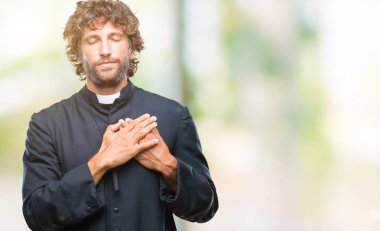 Yakışıklı İspanyol göğüs kapalı gözler ve yüzünde minnettar jest ile Katolik rahip adam ile gülümseyen izole arka plan üzerinde eller. Sağlık kavramı.