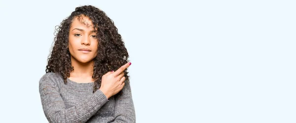 グレーを巻き毛を着ての若い美しい女性セーター広告 深刻な穏やかな顔を示す側面を手指で指す — ストック写真