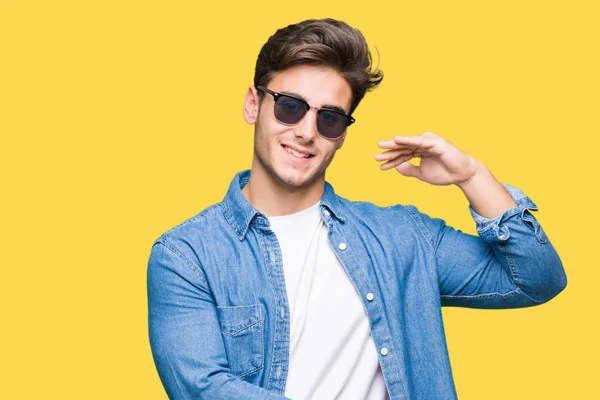 Young handsome man wearing sunglasses over isolated background gesturing with hands showing big and large size sign, measure symbol. Smiling looking at the camera. Measuring concept.