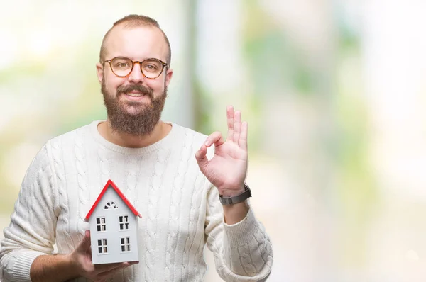 Young caucasian real state agent man holding house isolated background doing ok sign with fingers, excellent symbol