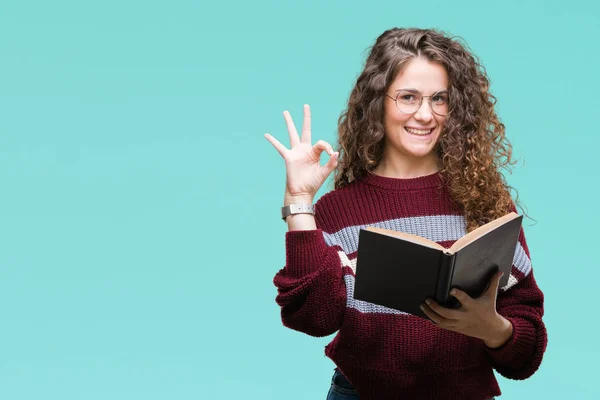 Young brunette girl reading a book wearing glasses over isolated background doing ok sign with fingers, excellent symbol