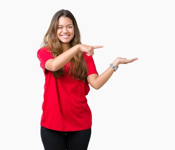 驚くし 手とを提示 指で指している間カメラに笑顔孤立の背景に赤い シャツを着て若い美しいブルネットの女性 — ストック写真