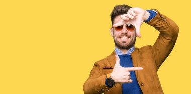 Genç yakışıklı zarif adam elleriyle yapım çerçeve ve parmakları ile mutlu yüz gülümseyen güneş gözlüğü takıyor. Yaratıcılık ve Fotoğrafçılık kavramı.