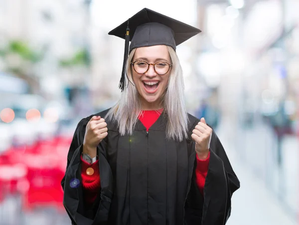 孤立した背景を祝う以上大学院制服を着て若いブロンドの女性は驚きし びっくりの腕の成功のため調達と開いた目 勝者の概念 — ストック写真